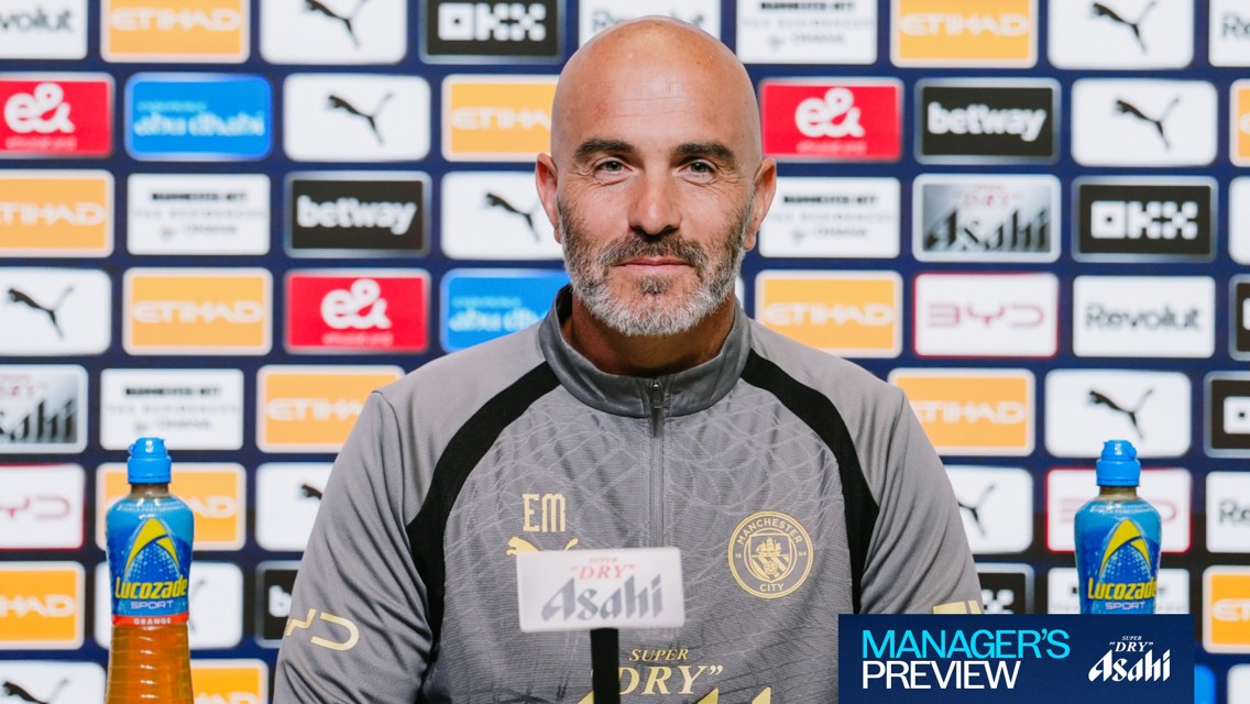 A bald man wearing a Manchester City jacket sits at a press conference table with sponsor logos like Etihad and Puma in the background.