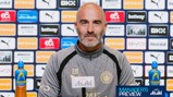 A bald man wearing a Manchester City jacket sits at a press conference table with sponsor logos like Etihad and Puma in the background.