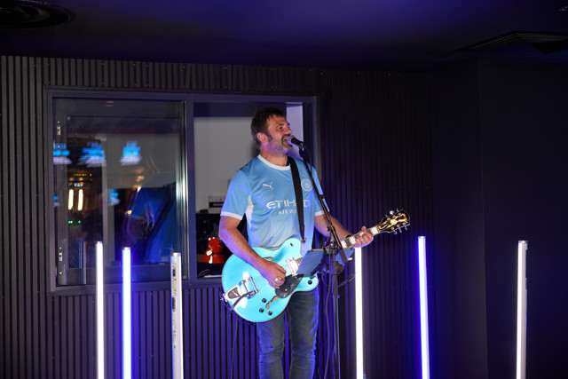 A person wearing a Manchester City jersey plays a blue electric guitar while singing into a microphone. Neon lights are visible in the background.