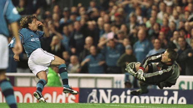 Soccer player in blue jersey attempts a goal, while opposing goalkeeper in gray dive-blocks the ball amidst a crowded stadium.