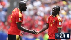 Two Belgium national team players shake hands on the field during a match. Both players are wearing red jerseys with yellow accents. A logo for Manchester City and FIFA World Cup is visible in the bottom corner.