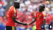 Two Belgium national team players shake hands on the field during a match. Both players are wearing red jerseys with yellow accents. A logo for Manchester City and FIFA World Cup is visible in the bottom corner.