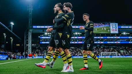 Three Manchester City players celebrating a goal on the field, with a scoreboard displaying 1-3 in the background.