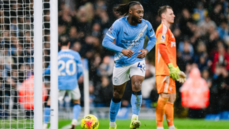 Manchester City player in action during a match, wearing the team's light blue kit. A goalkeeper is visible in orange attire.