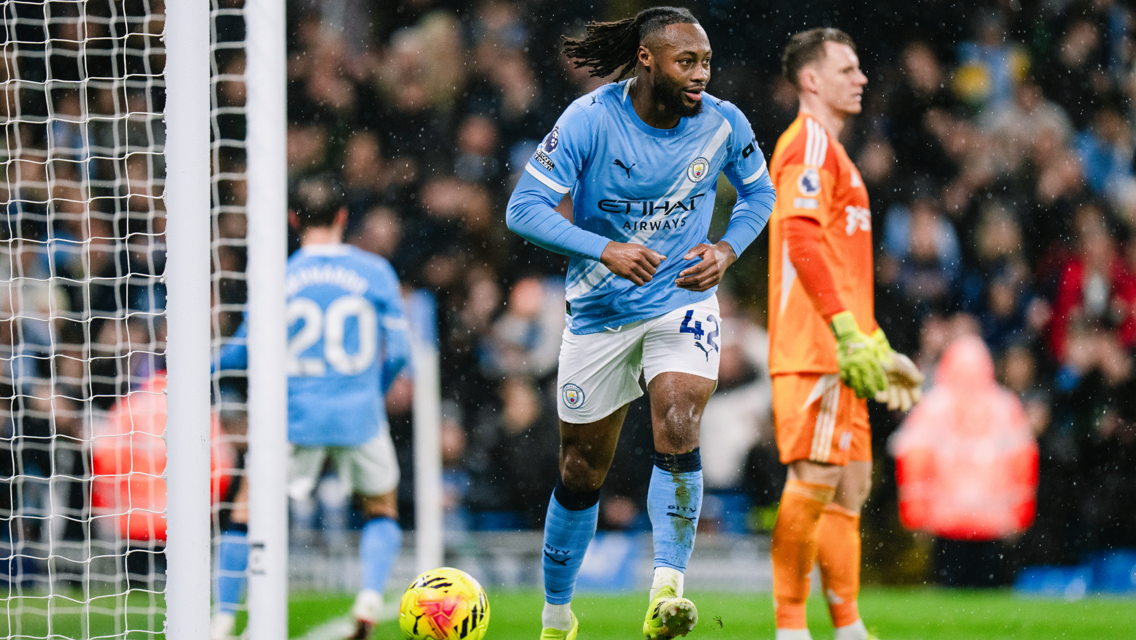 Manchester City player in action during a match, wearing the team's light blue kit. A goalkeeper is visible in orange attire.