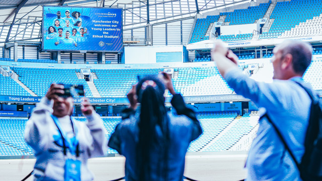 Visitors take photos at Etihad Stadium with a welcome message for Young Leaders on the screen. The stadium is empty with blue seats and a large welcome sign.