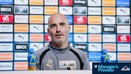 A person in a Manchester City jacket sits at a press conference table with Asahi branding, surrounded by a media backdrop featuring various sponsor logos.