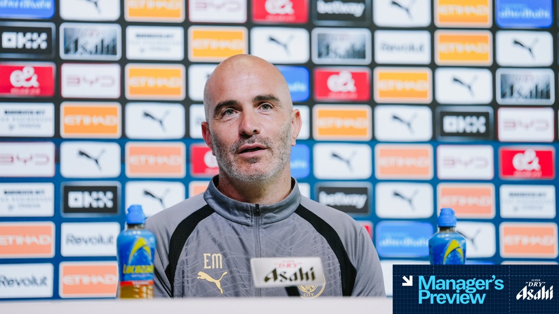 A person in a Manchester City jacket sits at a press conference table with Asahi branding, surrounded by a media backdrop featuring various sponsor logos.