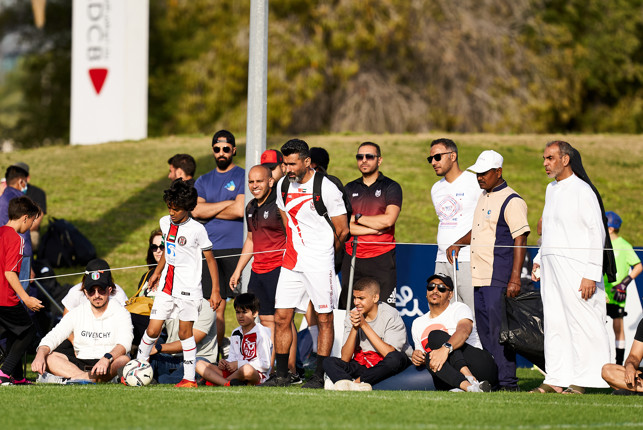 A group of adults and children standing and sitting near a football training session, watching young players on a grassy field.