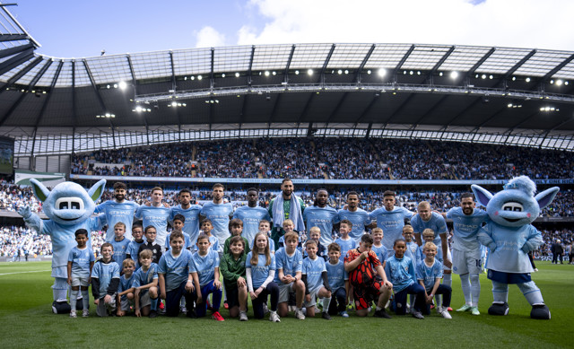 Group photo of Manchester City players and mascots on the field with young members at Etihad Stadium.