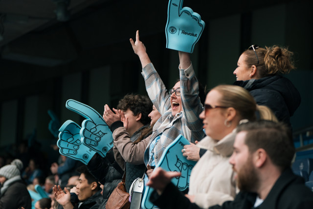Sports fans in a stadium enthusiastically hold large blue foam hands with the Revolut logo, cheering and clapping.