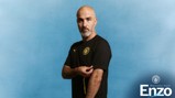 Man with a bald head and gray beard in a black shirt with a Manchester City logo stands against a blue background with 'Welcome back Enzo' text.