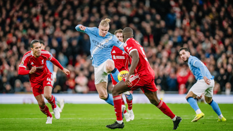 A soccer match with players from Manchester City and Liverpool competing for the ball. The player in blue is leaping over a player in red, with a crowd in the background.
