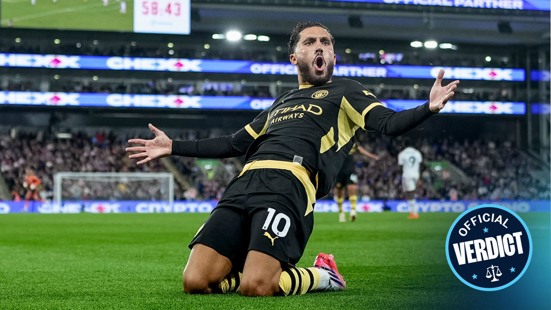 A football player wearing a black and yellow kit slides on his knees celebrating a goal, with a stadium crowd in the background. There is a digital scoreboard displaying 58:43.