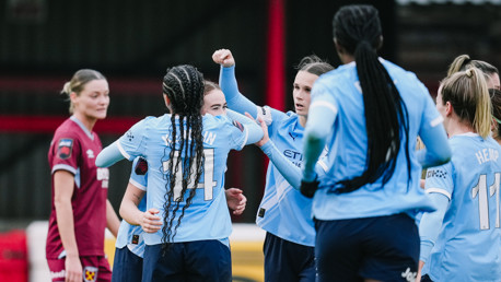 Women soccer players in light blue jerseys celebrate together on the field. One player in a maroon kit stands nearby.