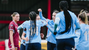 Women soccer players in light blue jerseys celebrate together on the field. One player in a maroon kit stands nearby.