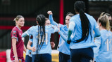 Women soccer players in light blue jerseys celebrate together on the field. One player in a maroon kit stands nearby.