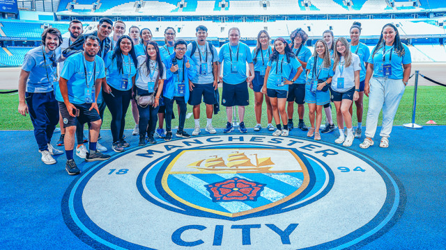 A group of people wearing Man City lanyards and t-shirts posing together inside Etihad Stadium standing on the Manchester City logo on the floor.