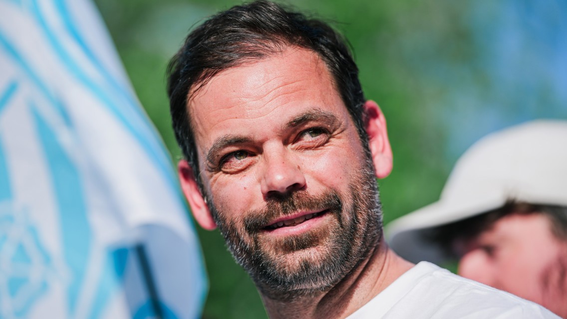 A man with a beard and short dark hair is smiling outdoors with a blurred background featuring a flag.
