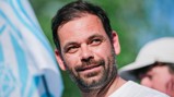 A man with a beard and short dark hair is smiling outdoors with a blurred background featuring a flag.
