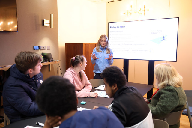 A woman in blue presents on 'Plan, Lead, and Evaluate' to a group seated around a table, with a screen displaying presentation details.