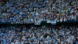 A large crowd of Manchester City fans at Etihad Stadium waving blue and white flags.