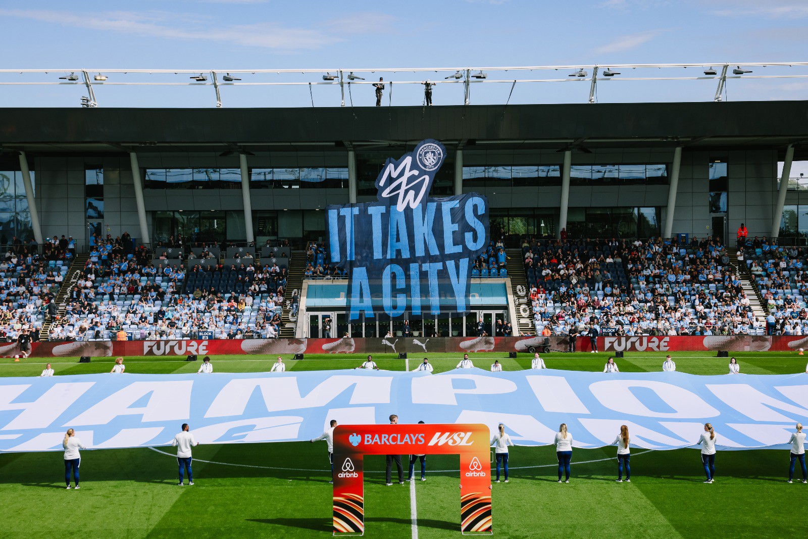 A large banner saying 'IT TAKES A CITY' is displayed in the stands during a Manchester City women's game at the Academy Stadium. The field shows a large sign with 'CHAMPIONS' and the Barclays Women's Super League branding.