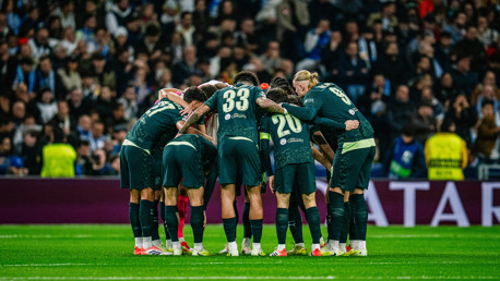 Football team huddle on the field with players in dark jerseys and numbers visible. Crowd in background.