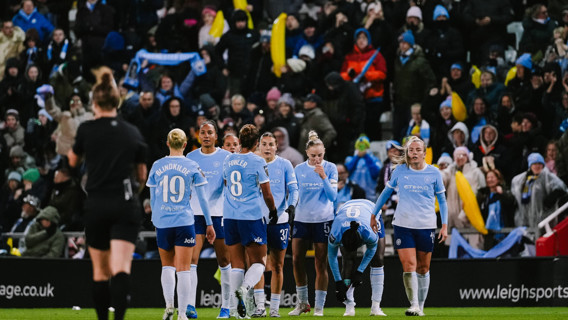 Manchester City women's football team in blue uniforms celebrating on the field with fans in the background holding scarves and inflatables.