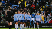 Manchester City women's football team in blue uniforms celebrating on the field with fans in the background holding scarves and inflatables.