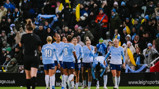 Manchester City women's football team in blue uniforms celebrating on the field with fans in the background holding scarves and inflatables.