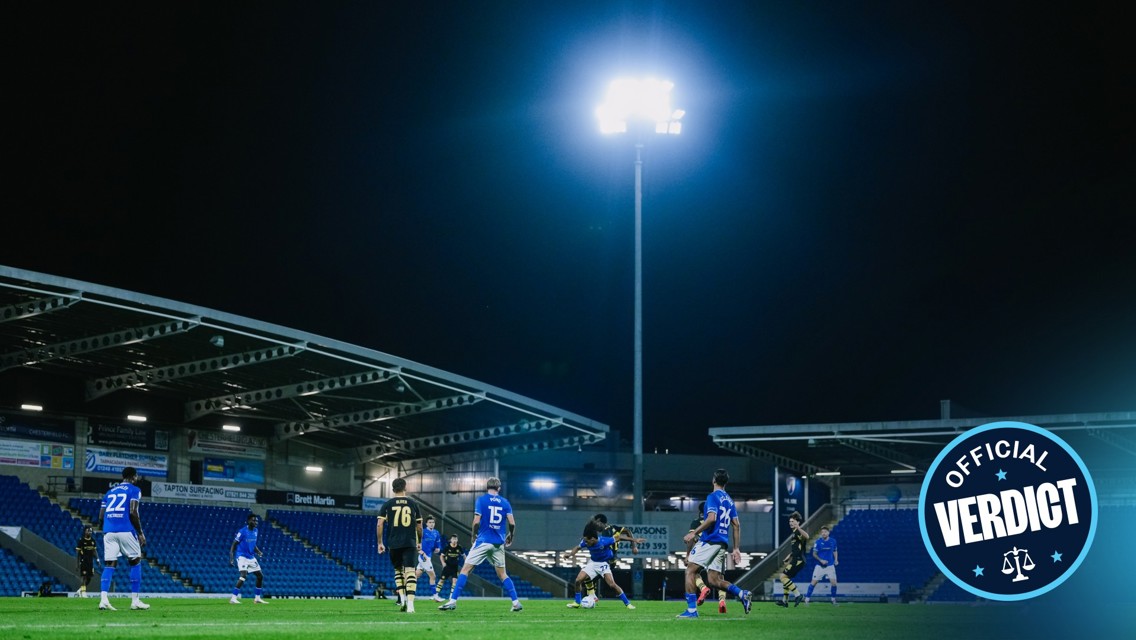 Nighttime football match with players in blue jerseys and bright floodlights at a stadium with empty stands.