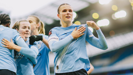 Manchester City women's team players celebrate on the pitch, with one player holding the shirt proudly at the stadium.