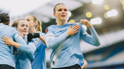 Manchester City women's team players celebrate on the pitch, with one player holding the shirt proudly at the stadium.
