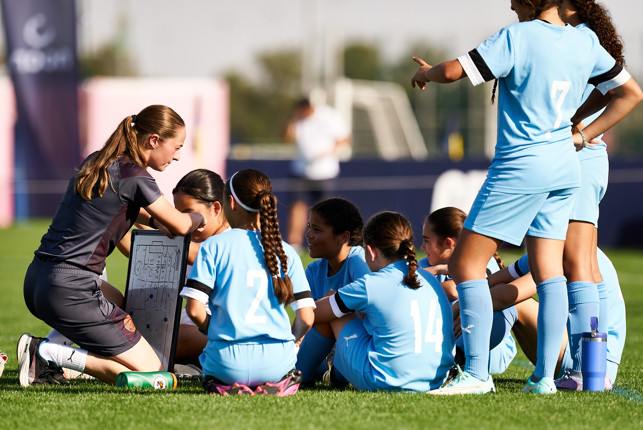 A woman coach holds a strategy board while talking to a Manchester City Women's Team in light blue uniforms during a training session on a soccer field.