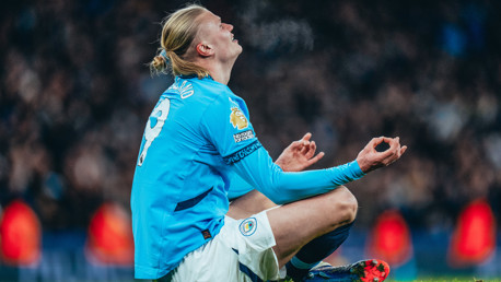 A soccer player wearing a Manchester City jersey, number 9, sits on the field in a meditative pose during a game. The Premier League logo is visible on the sleeve.