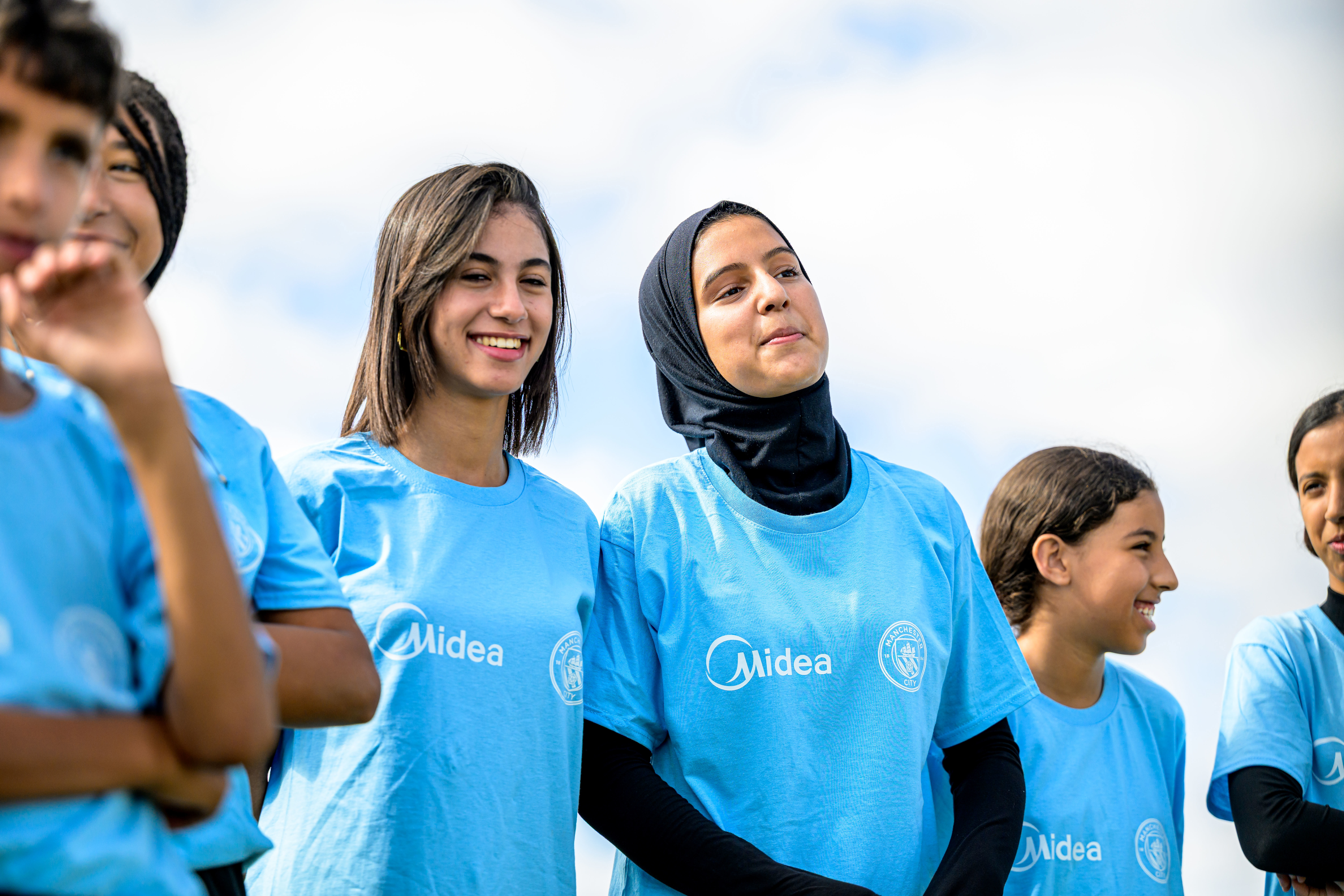 A group of young people wearing light blue Midea-branded shirts, standing outdoors with a clear sky background.