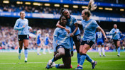 Group of Manchester City women football players celebrating on the field. One player is kneeling with others hugging and celebrating together.