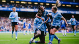 Group of Manchester City women football players celebrating on the field. One player is kneeling with others hugging and celebrating together.