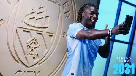 A person wearing a light blue Manchester City jersey taking a selfie in front of the Manchester City emblem. The text 'JEREMY DOKU 2031' is displayed.