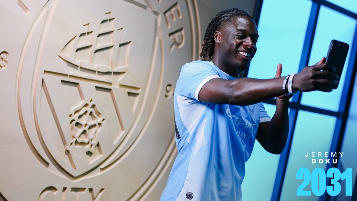 A person wearing a light blue Manchester City jersey taking a selfie in front of the Manchester City emblem. The text 'JEREMY DOKU 2031' is displayed.
