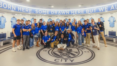 Group wearing blue shirts posing in Manchester City dressing room with player jerseys in background.