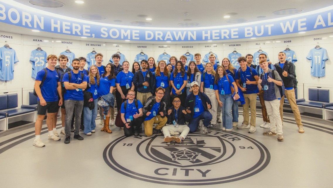 Group wearing blue shirts posing in Manchester City dressing room with player jerseys in background.