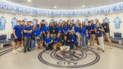 Group wearing blue shirts posing in Manchester City dressing room with player jerseys in background.