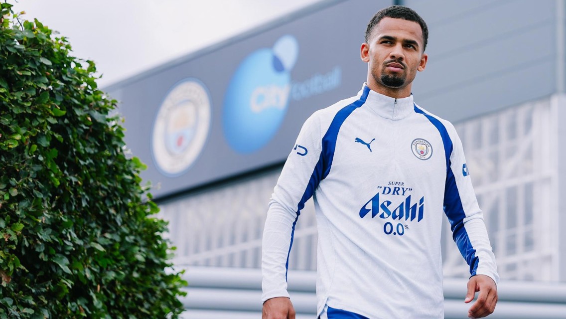 A person wearing a Manchester City training tracksuit walks outside the City Football Academy. The tracksuit features Puma branding and an Asahi Super Dry logo.