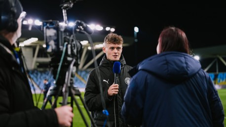 A person in a Manchester City jacket is interviewed on a football field at night with bright floodlights. A camera and microphone are visible.