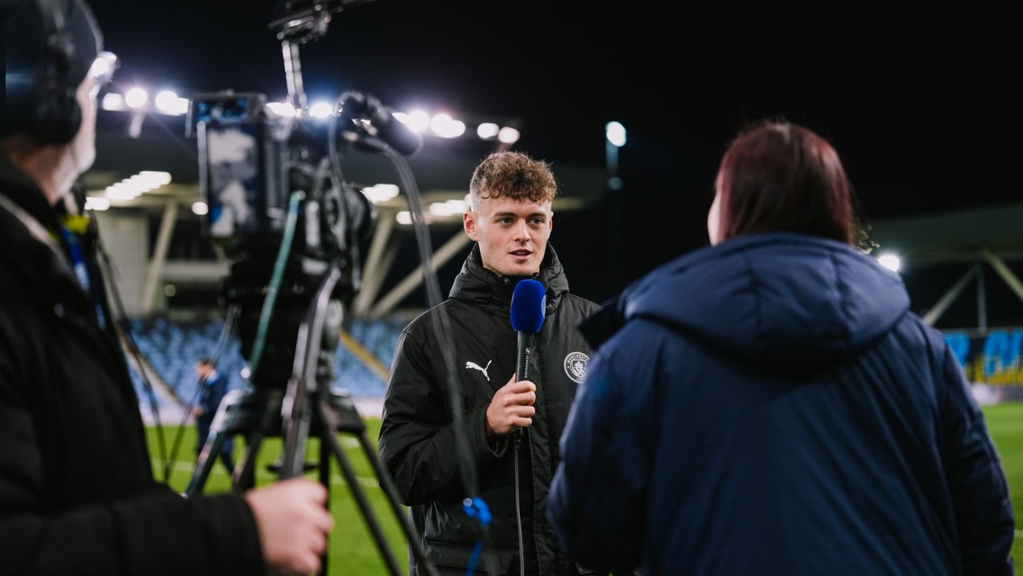 A person in a Manchester City jacket is interviewed on a football field at night with bright floodlights. A camera and microphone are visible.