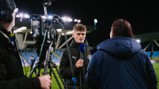 A person in a Manchester City jacket is interviewed on a football field at night with bright floodlights. A camera and microphone are visible.