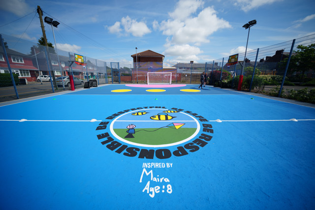 An outdoor basketball court with colorful floor markings, including bees, circles, and a kite. The court's design is inspired by Maira, Age 8, with a slogan encouraging respect, safety, and responsibility. There are houses and a small building in the background.