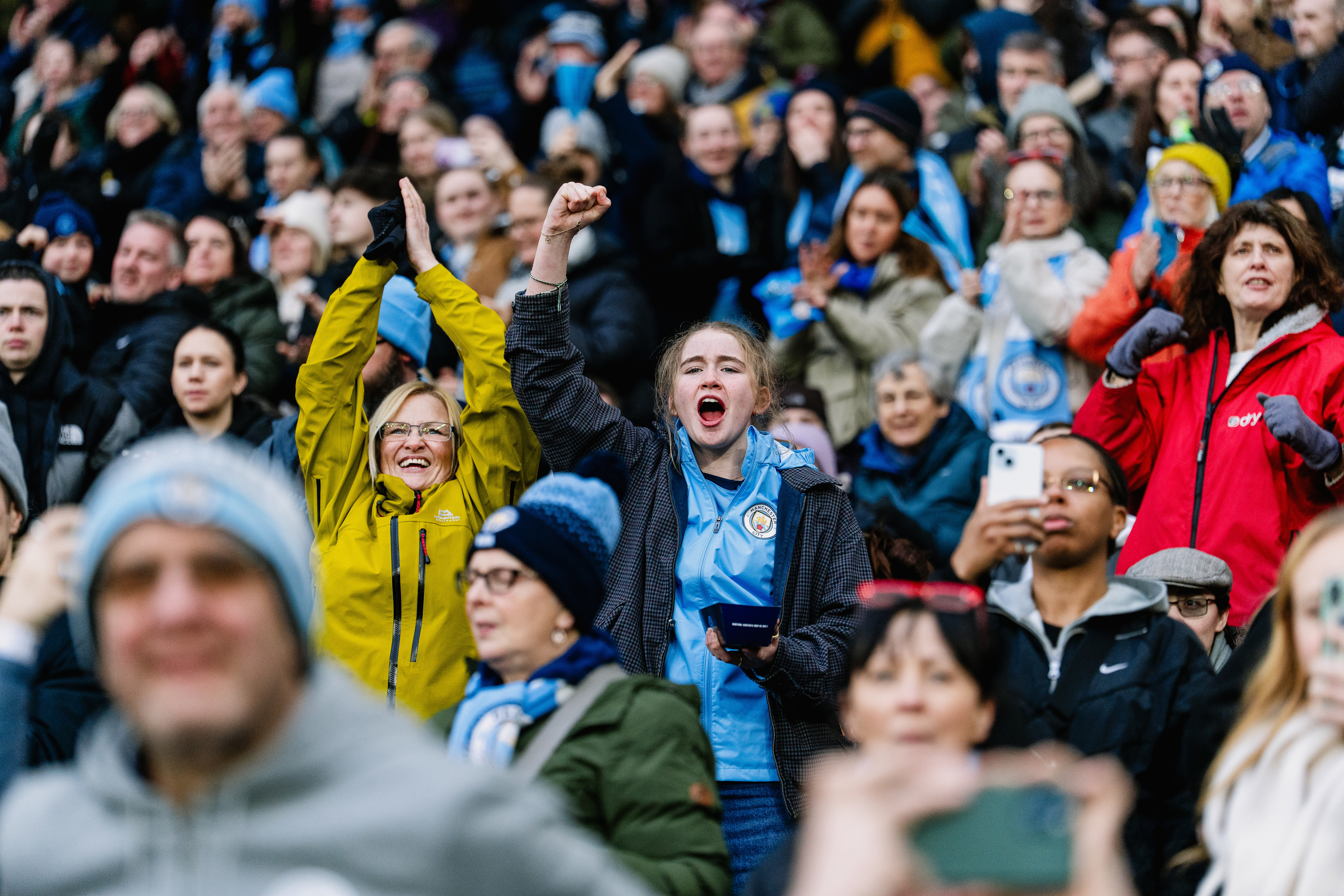 Crowd of Manchester City fans celebrating in a stadium, with visible team logos on clothing.
